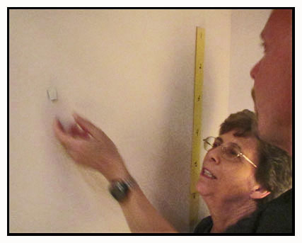 Photo shows Dona reaching for a piece of paper taped to the wall.  The paper is almost the same color as the wall and is very hard to see.